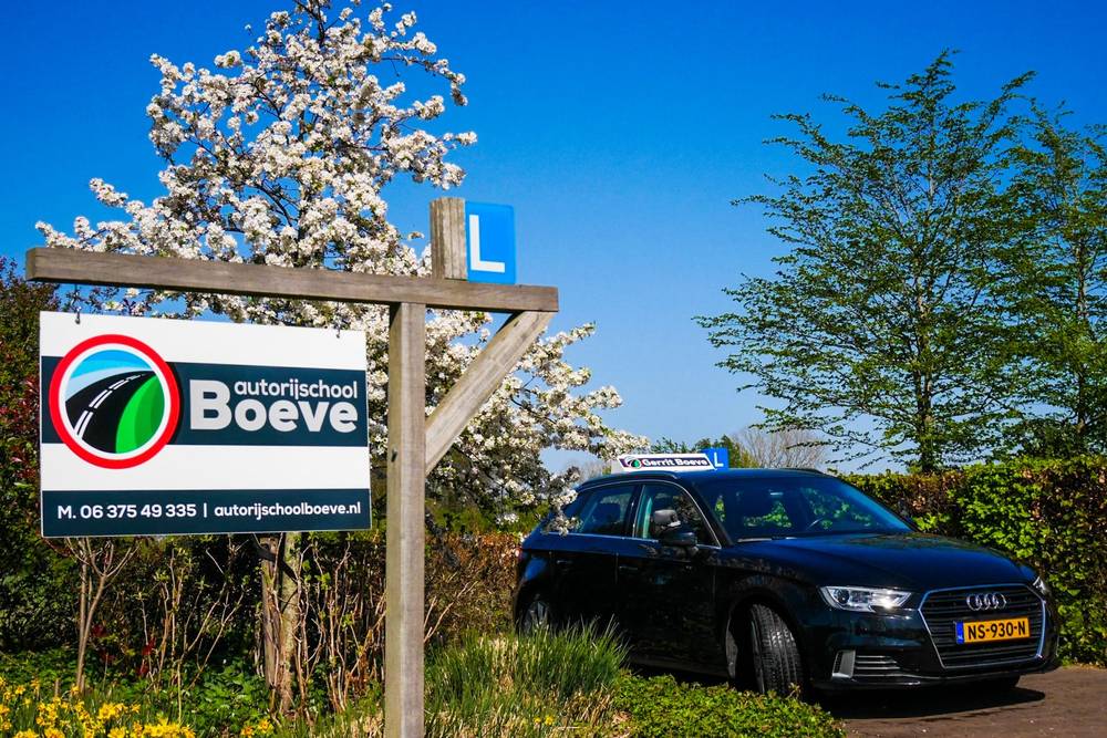 Het reclamebord van Autorijschool Boeve langs de weg in Sint Jansklooster, met een bloeiende boom en de lesauto erachter